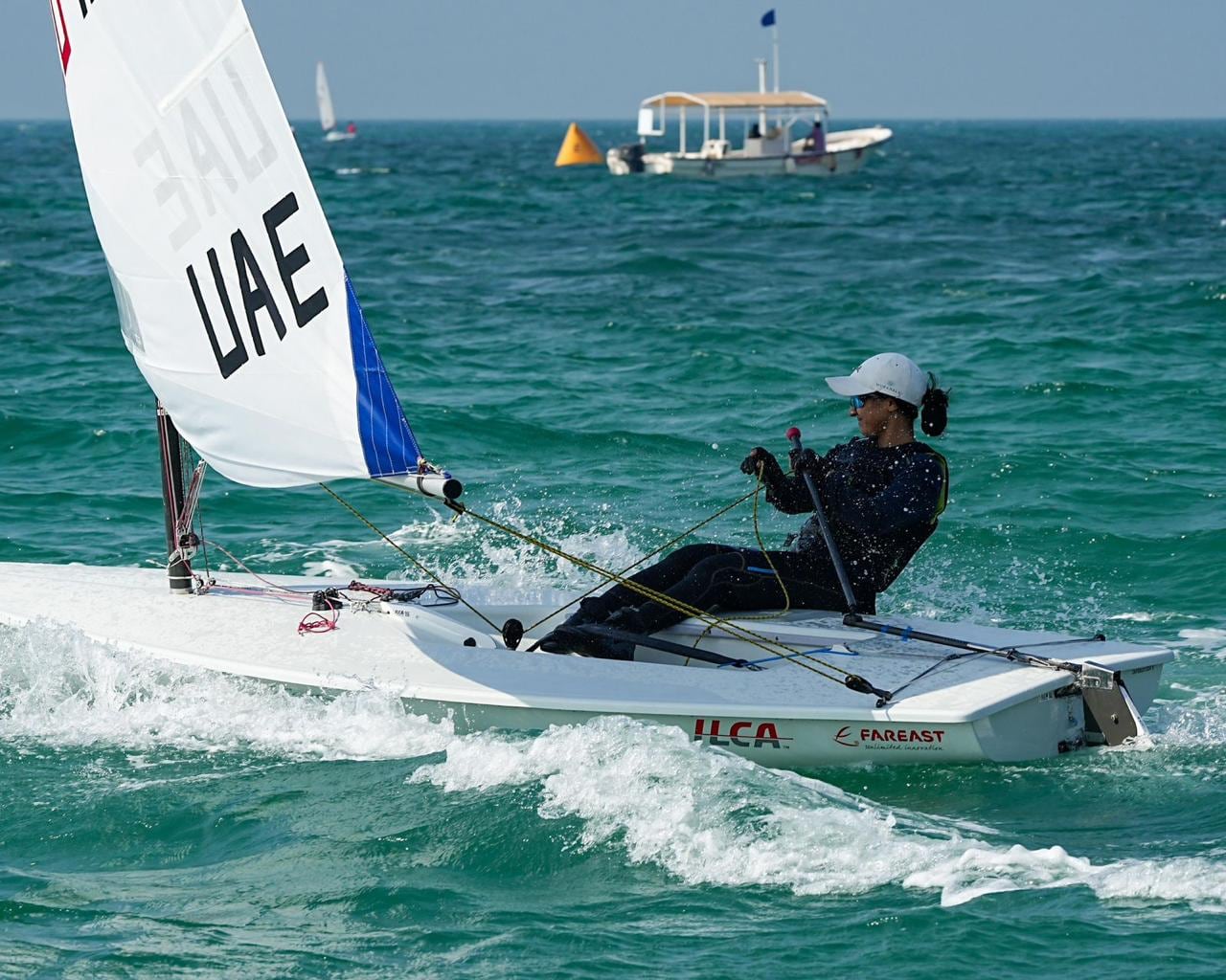 A sailor hiking out on an ILCA dinghy under a sail marked UAE, with a coach boat behind.